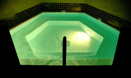 Ein sechseckiger Pool mit einer umlaufenden Stufe ist mit einer Unterwasserbeleuchtung ausgestattet, die das Wasser im Dunklen türkis wirken lässt.