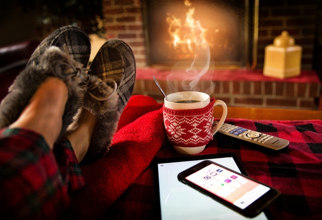 Jemand sitzt mit dicken Hausschuhen gemütlich vor dem Kamin und einer Tasse Tee, daneben liegt ein Handy, mit dem man die Helligkeit des Lichtes steuern könnte.