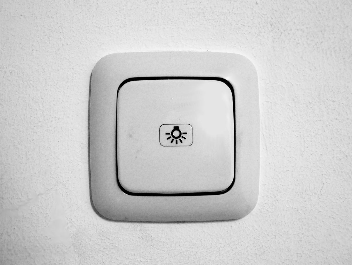 An einer weißen Wand sieht man einen weißen, einfach Lichtschalter, der ein Glühlampen-Symbol mittig aufgebracht trägt.