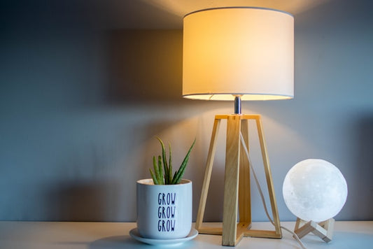 Eine Tischleuchte steht auf einer Holzoberfläche. Sie strahlt warmes Licht ab. Neben ihr steht ein kleiner Blumentopf mit Grünpflanze darin.