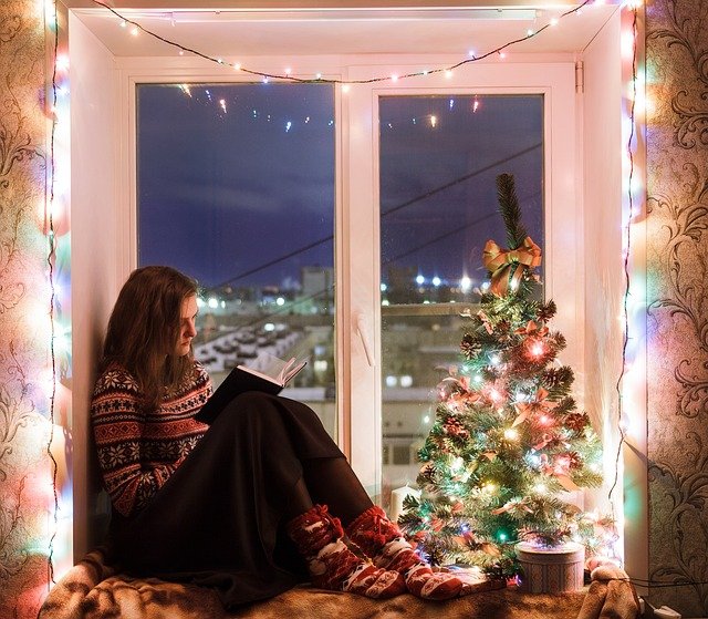 Eine junge Frau sitzt lesend auf einem Fensterbrett, welches mit vielen bunten Weihnachtslichtern geschmückt ist.